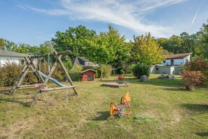 Ein Spielplatz mit Schaukeln und Rutschen, umgeben von Bäumen und einem kleinen, roten Spielhäuschen.