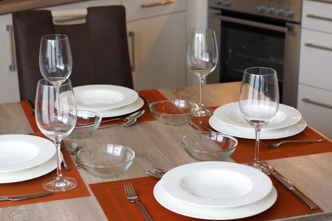Une table à manger dressée avec de l’assiette blanche, des bols en verre transparent et des verres à vin, agrémentée d’un chemin de table en bois et d’une chaise marron en arrière-plan.