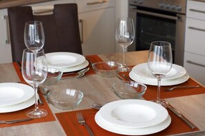 Une table à manger dressée avec de l’assiette blanche, des bols en verre transparent et des verres à vin, agrémentée d’un chemin de table en bois et d’une chaise marron en arrière-plan.