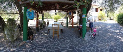 A covered outdoor seating area with a table and chairs, surrounded by greenery and a stone-paved path.