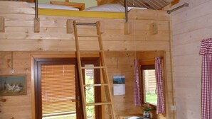 Interior de una cabaña de madera con cama en el altillo, escalera de madera, zona de comedor con mesa y sillas, y ventana con cortinas.