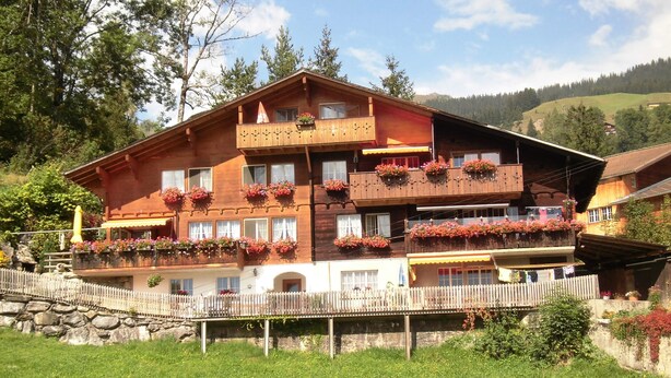 Un immeuble en bois à plusieurs étages, doté de balcons ornés de jardinières fleuries, se dressant sur un fond de paysage montagneux.