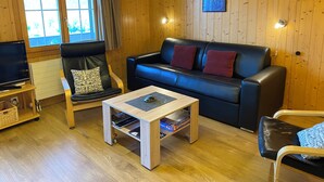 Et rum med træpaneler, en sort lædersofa, et træbord, et fjernsyn og et vindue med gardiner.