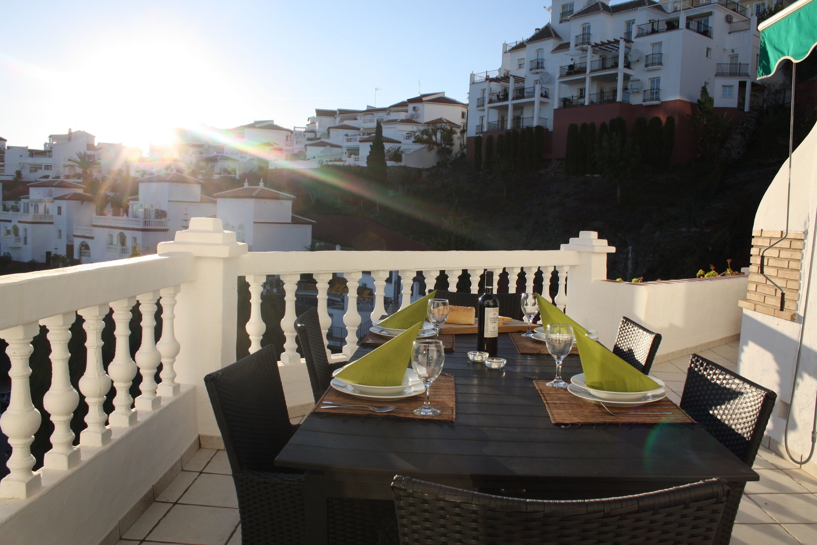 A dining table set with plates, glasses, and a bottle, overlooking a residential area with white buildings and a clear sky.