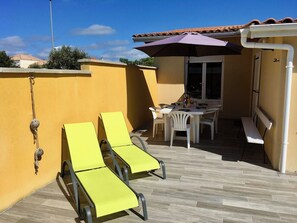 Eine sonnige Terrassenanlage mit zwei gelben Liegestühlen, einem Tisch mit Stühlen und einem violetten Sonnenschirm.