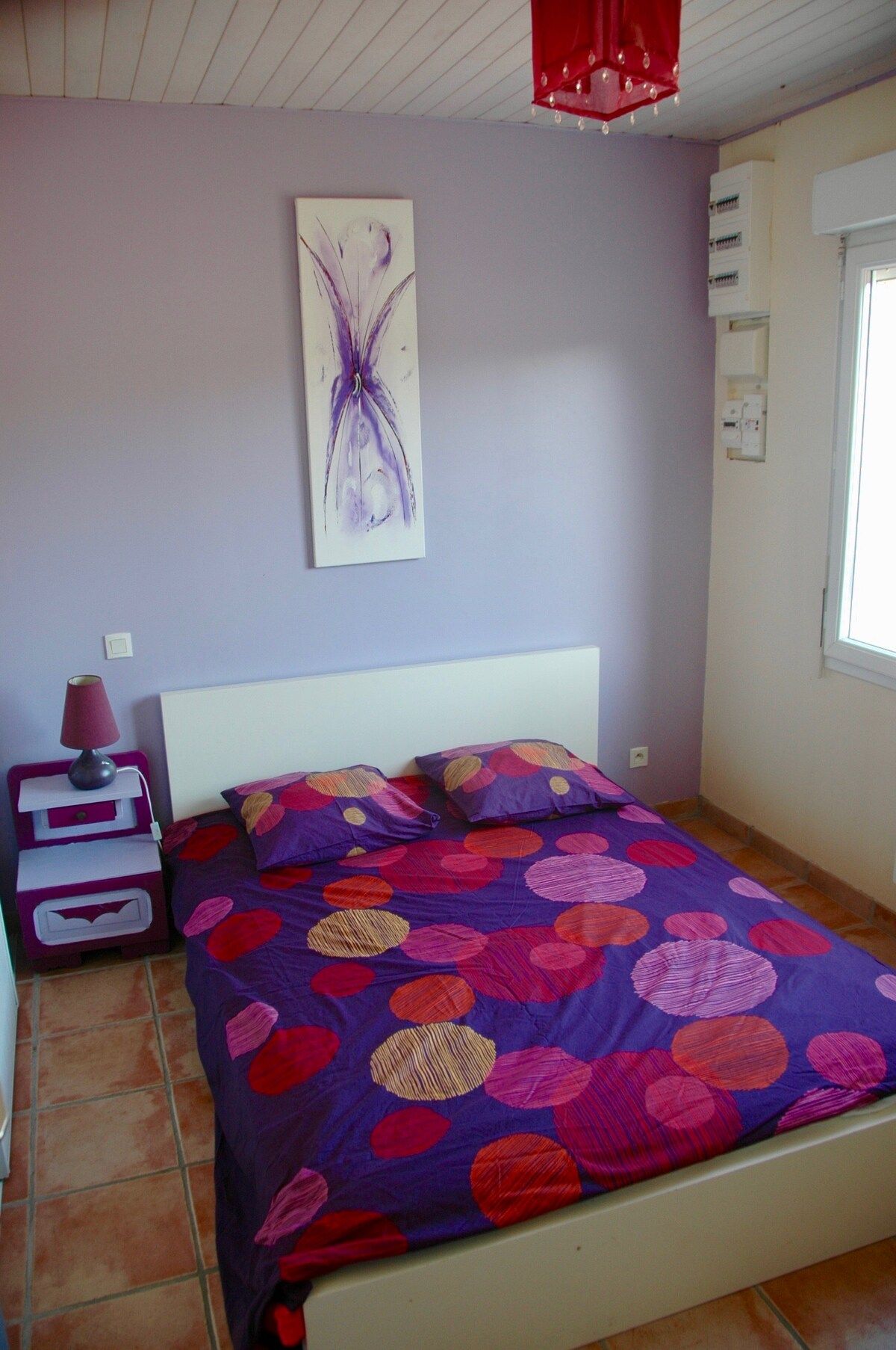 Une chambre à coucher comprenant un lit, une table de chevet, une lampe, un tableau décoratif et une fenêtre.