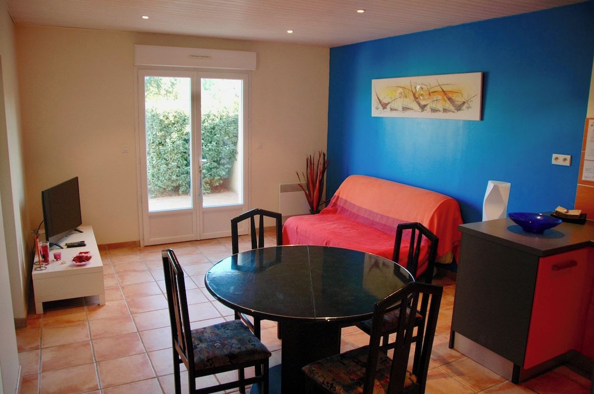 A room with a bed, a dining table with chairs, a television on a stand, and a window with a view of greenery.