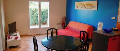 A room with a bed, a dining table with chairs, a television on a stand, and a window with a view of greenery.