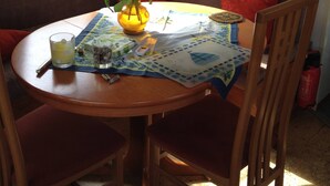 Una mesa redonda de madera con un florero amarillo de tulipanes, una vela y un mantel individual con temática náutica. Alrededor de la mesa hay sillas de madera y un cojín rojo en el banco detrás.