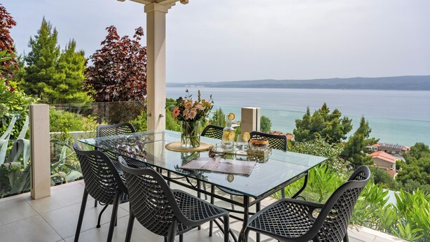 Ein Tisch mit Glasplatte, Stühle, eine Vase mit Blumen und Blick auf das Meer.