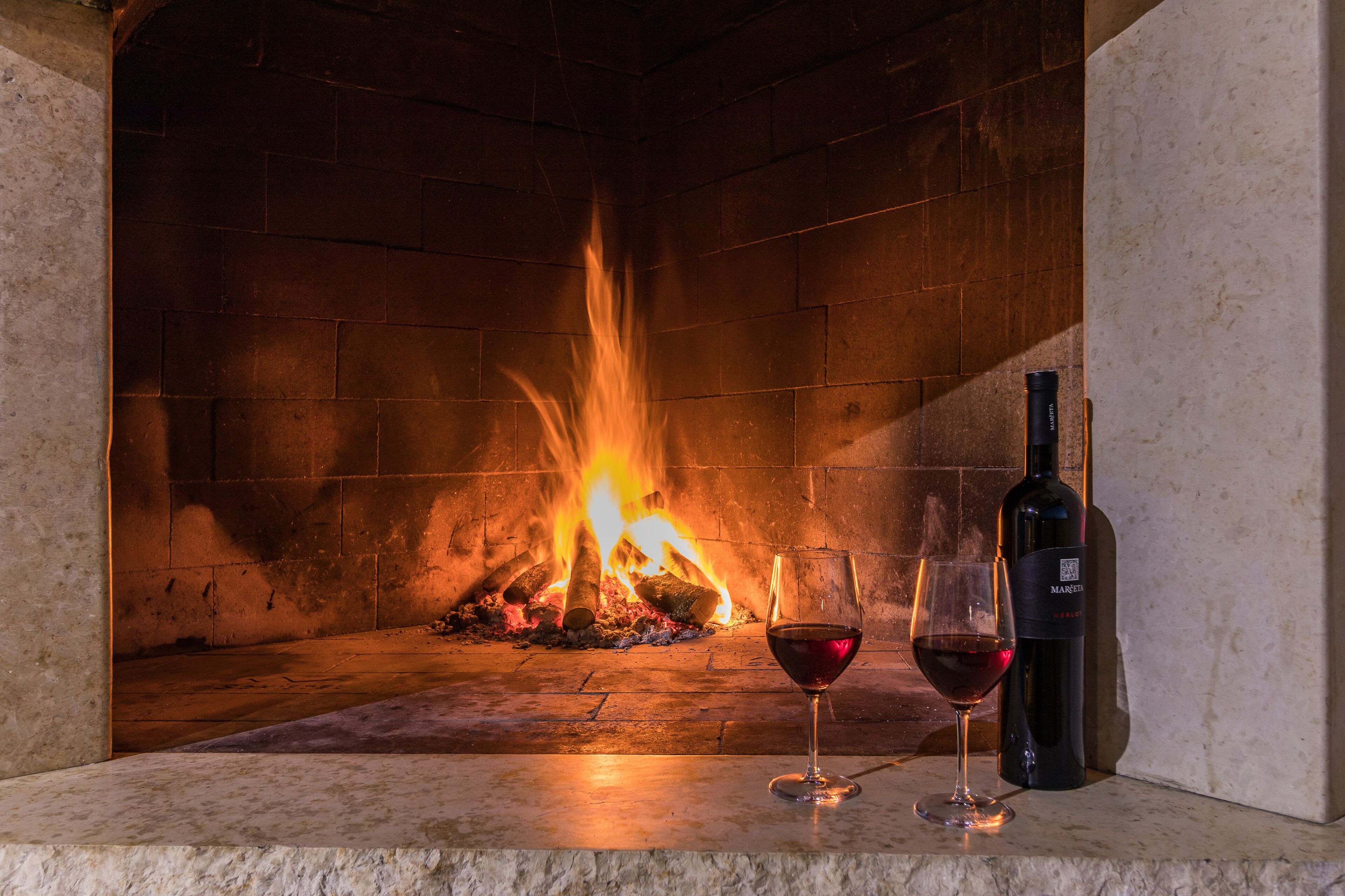 A fireplace with burning logs, a bottle of wine, and two wine glasses on a stone mantel.
