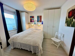 A bedroom with a bed, a white chair, a wardrobe, a ceiling light, and artwork on the wall. - Apartment only 600 m. from the beach, with sunny balcony on the south! (Nordholland)