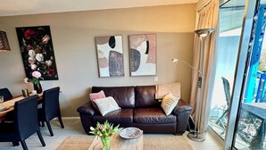 A modern living room with a leather sofa, a wooden coffee table, and a dining area with chairs. There are abstract paintings on the wall and a large window with a view of the outdoors. - Apartment only 600 m. from the beach, with sunny balcony on the south! (Nordholland)