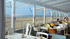 A restaurant with a view of the beach, featuring white tables and chairs, and a glass ceiling. - Apartment only 600 m. from the beach, with sunny balcony on the south! (Nordholland)