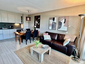 A modern living room with a dark leather sofa, a wooden coffee table, and a dining area with a wooden table and chairs. There are abstract paintings on the wall and a floor lamp. - Apartment only 600 m. from the beach, with sunny balcony on the south! (Nordholland)