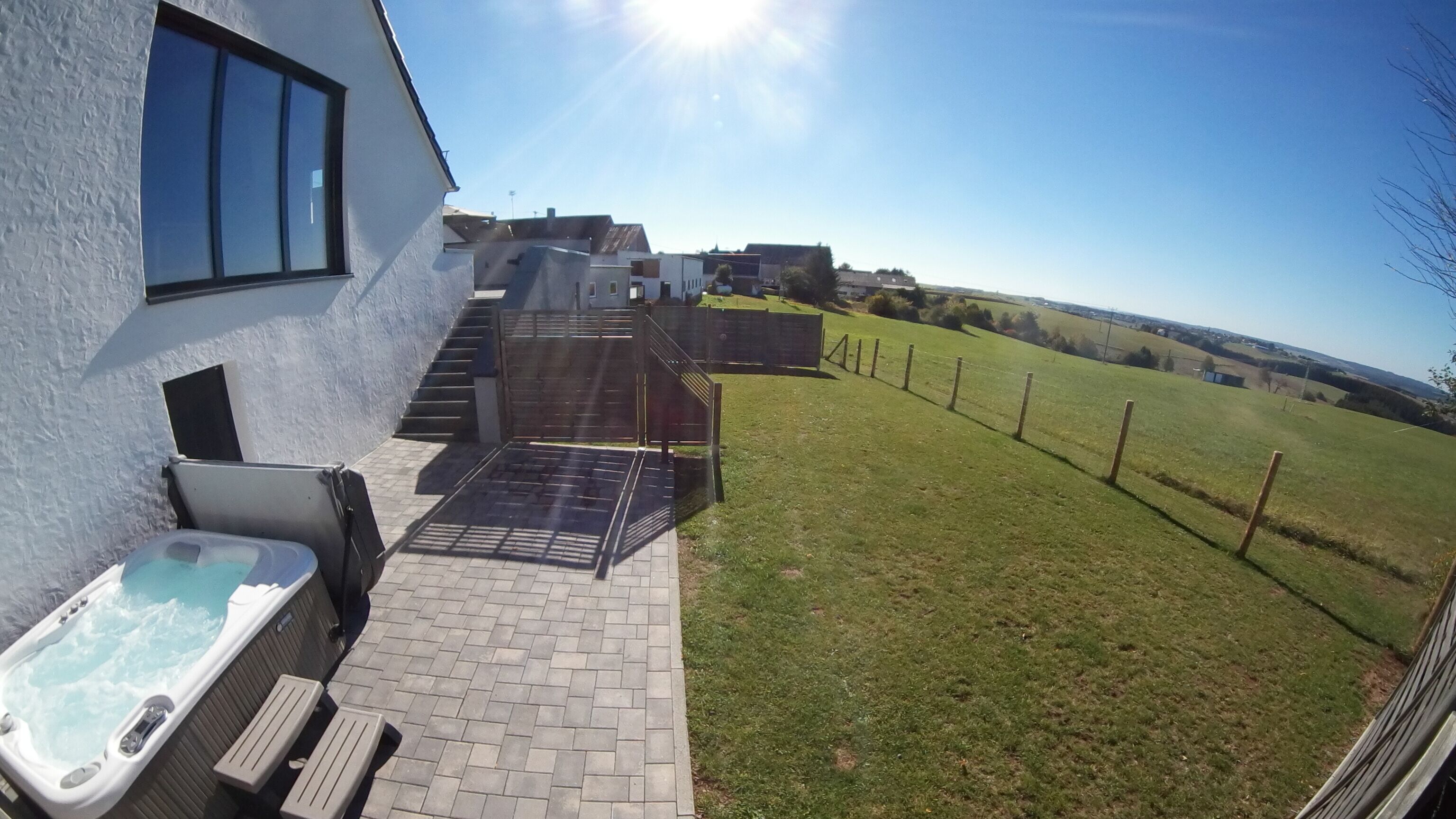 A patio with a hot tub, steps leading to a gated area, and a view of a grassy field and distant buildings.