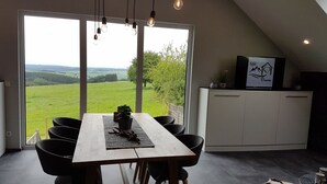 A dining area with a table and chairs, a view of a green landscape through large windows, a cabinet with a TV, and a ceiling with hanging lights.