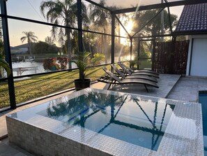 Una piscina interior con borde de azulejos, sillones de descanso y vistas a palmeras y un cuerpo de agua en el exterior.