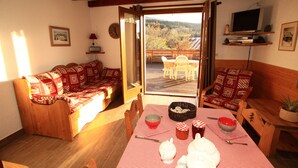 Uma sala de estar acolhedora com um sofá de madeira, uma mesa de jantar com toalha quadriculada e uma varanda com vista para uma paisagem montanhosa.