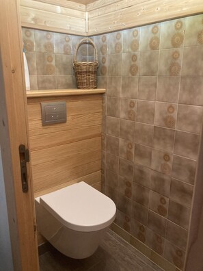 A small bathroom with a toilet, a wooden shelf with a basket, and patterned tiles. - Comfortable and well-kept Vercors house, made beds, closed garage & Wifi (Villard-De-Lans)
