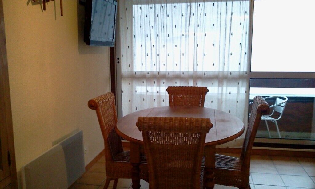 Un comedor con una mesa redonda y cuatro sillas de mimbre, una ventana con cortinas estampadas y un televisor fijado en la pared.