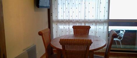 Un comedor con una mesa redonda y cuatro sillas de mimbre, una ventana con cortinas estampadas y un televisor fijado en la pared.