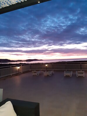 A balcony with white railings and lounge chairs, overlooking a body of water with distant land and a sunset sky.