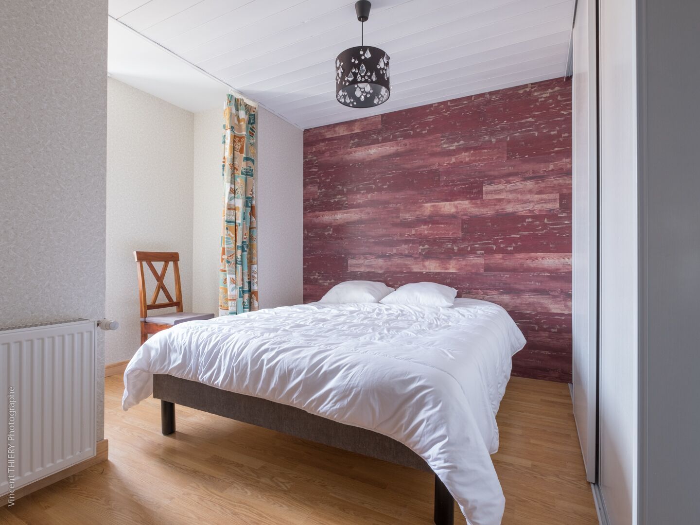 A bedroom with a wooden floor, a bed with white bedding, a wooden chair, a radiator, and a patterned curtain.