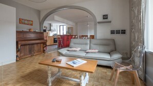 Een woonkamer met een houten salontafel, een grijze bank en een transparante stoel. Achterin staat een houten kast en een piano.