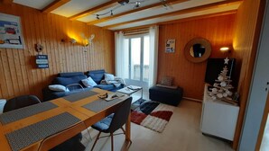 A cozy room with a wooden wall, a blue sofa, a dining table with chairs, a small black sofa, and a TV on a stand. - Rent cosy apartment in the heart of Lioran resort close to the slopes (Laveissière)