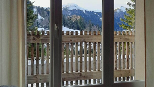 A room with a large sliding glass door leading to a balcony with a wooden railing and a view of snow-capped mountains. - Rent cosy apartment in the heart of Lioran resort close to the slopes (Laveissière)