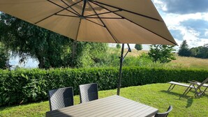 An outdoor patio with a table and chairs under a large umbrella, surrounded by greenery and a view of a lake. - Idyllic natural oasis - pure tranquillity just 5 minutes' walk from the Müritz  (Ludorf)