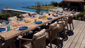 Eine Holzterrasse mit einem Esstisch, gedeckt für sechs Personen, mit Blick auf einen Pool und das Meer.