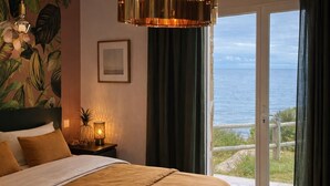 Ett sovrum med en stor säng, ett nattduksbord, en lampa, en inramad tavla och utsikt över havet genom en skjutdörr.