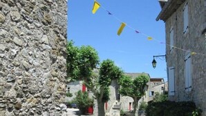 A stone building with a balcony, a street with parked cars, and a clear blue sky. - Cozy cottage in the village of 4 Tours (Valvignères)