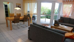 Una sala de estar con un sofá gris, una mesa de comedor con sillas de mimbre y un balcón con vistas a zonas verdes.