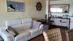 A living room with a white sofa, a small table, a wicker chair, a ship's wheel, and a framed picture on the wall. - YOU Escape IN BAY OF Roses, ONE OF THE Most Beautiful World! (EMPURIABRAVA)