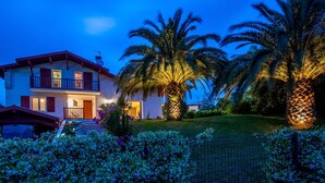 A two-story house with a balcony, surrounded by well-maintained gardens and palm trees.