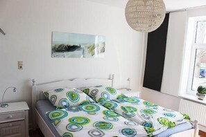 A bedroom with a bed, a bedside table, a lamp, a hanging light fixture, a window, and a wall-mounted picture.