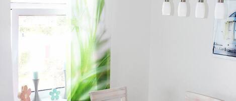 Un coin repas avec une table et des chaises blanches, un luminaire suspendu et une fenêtre agrémentée d’une plante.