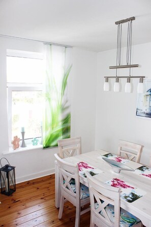 A dining area with a white table and chairs, a hanging light fixture, and a window with a plant.
