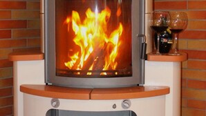 Een houtkachel met een glazen ruit, een opbergruimte voor hout eronder, en een bakstenen muur op de achtergrond.