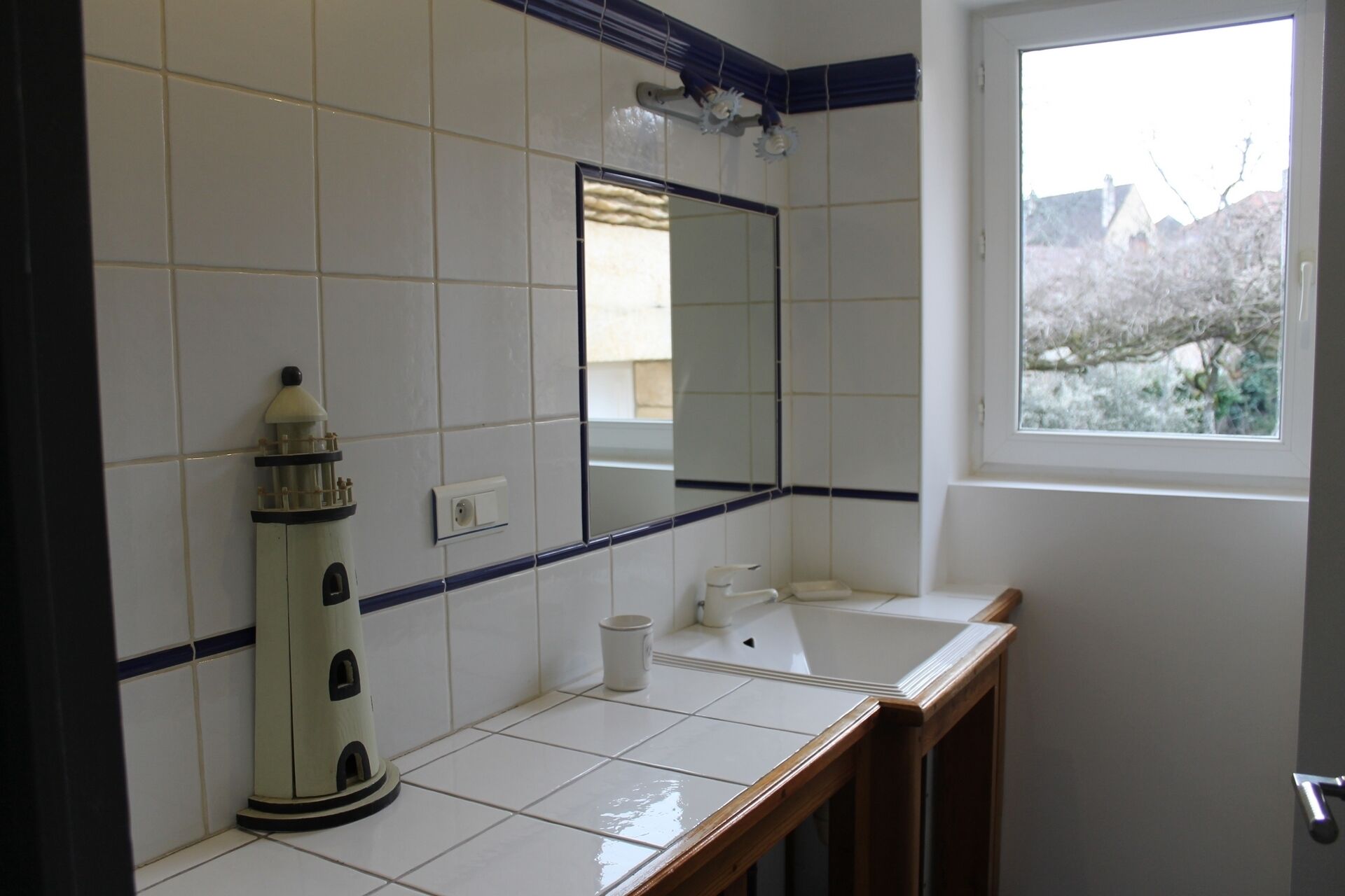 A bathroom with white tiles, a mirror, a window, and a lighthouse-shaped soap dispenser.