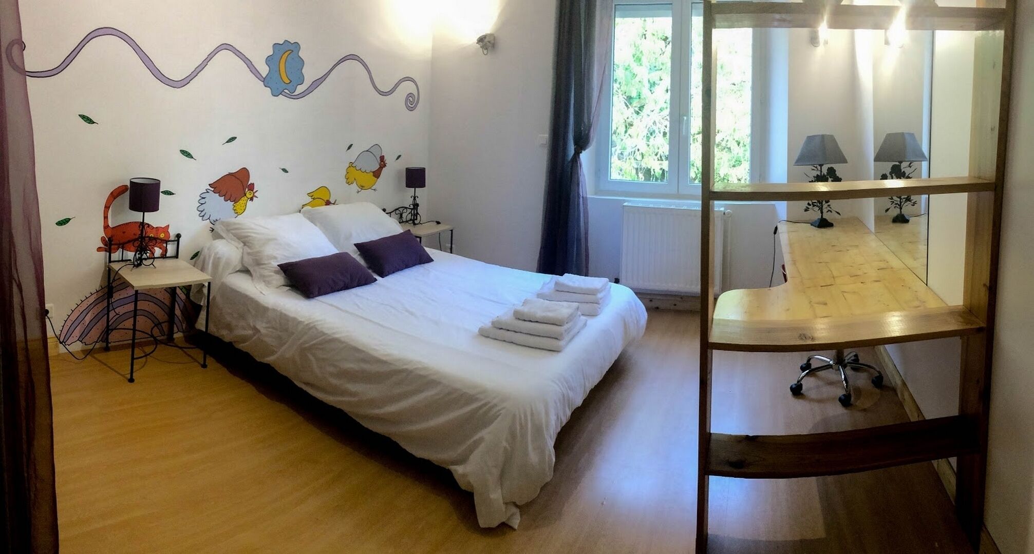 A neatly made bed with white linens and purple pillows, a small table with a lamp, and a wooden desk with two lamps.