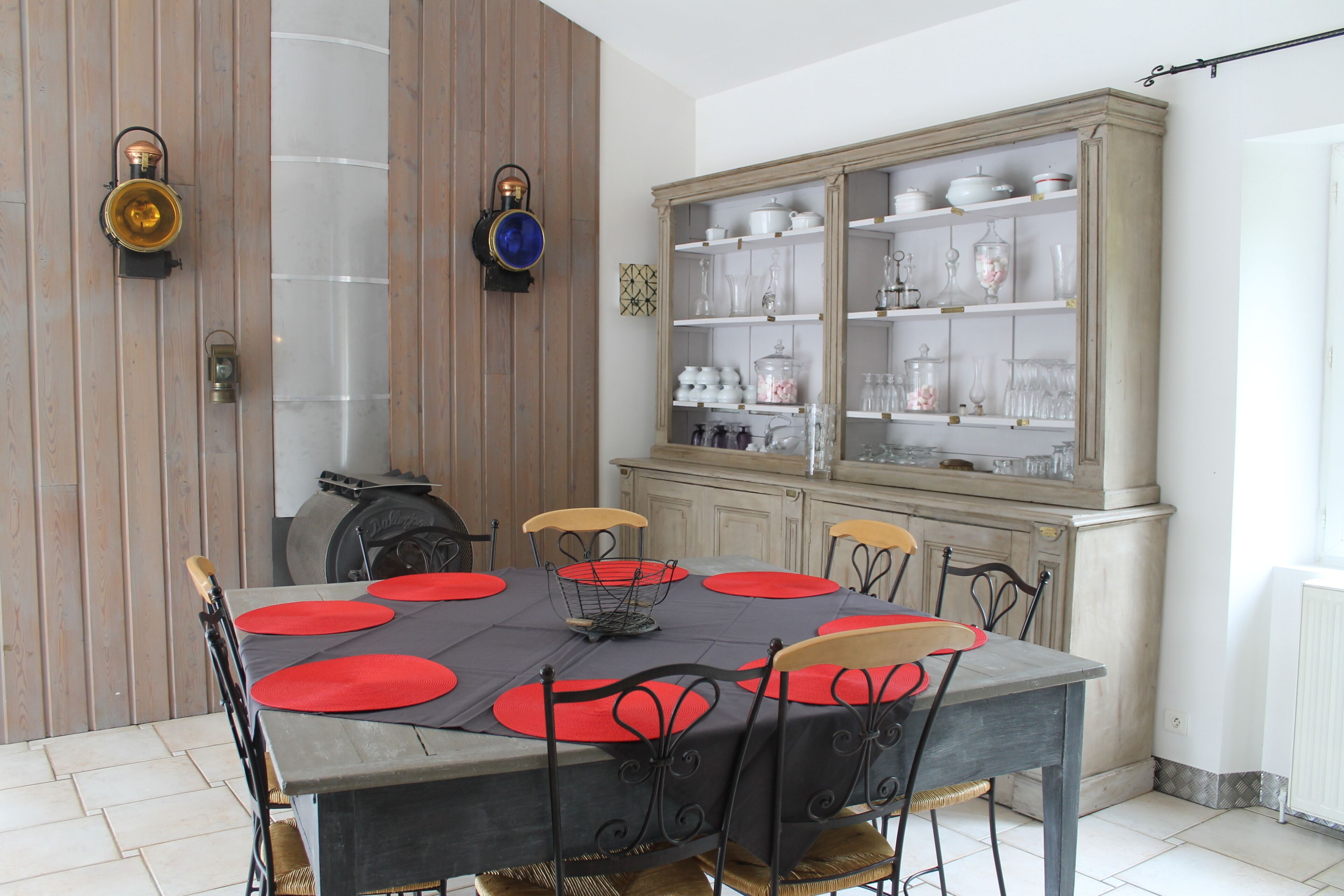 A dining area with a table set for six, surrounded by six chairs with red cushions. A cabinet with glassware and dishes is to the right.