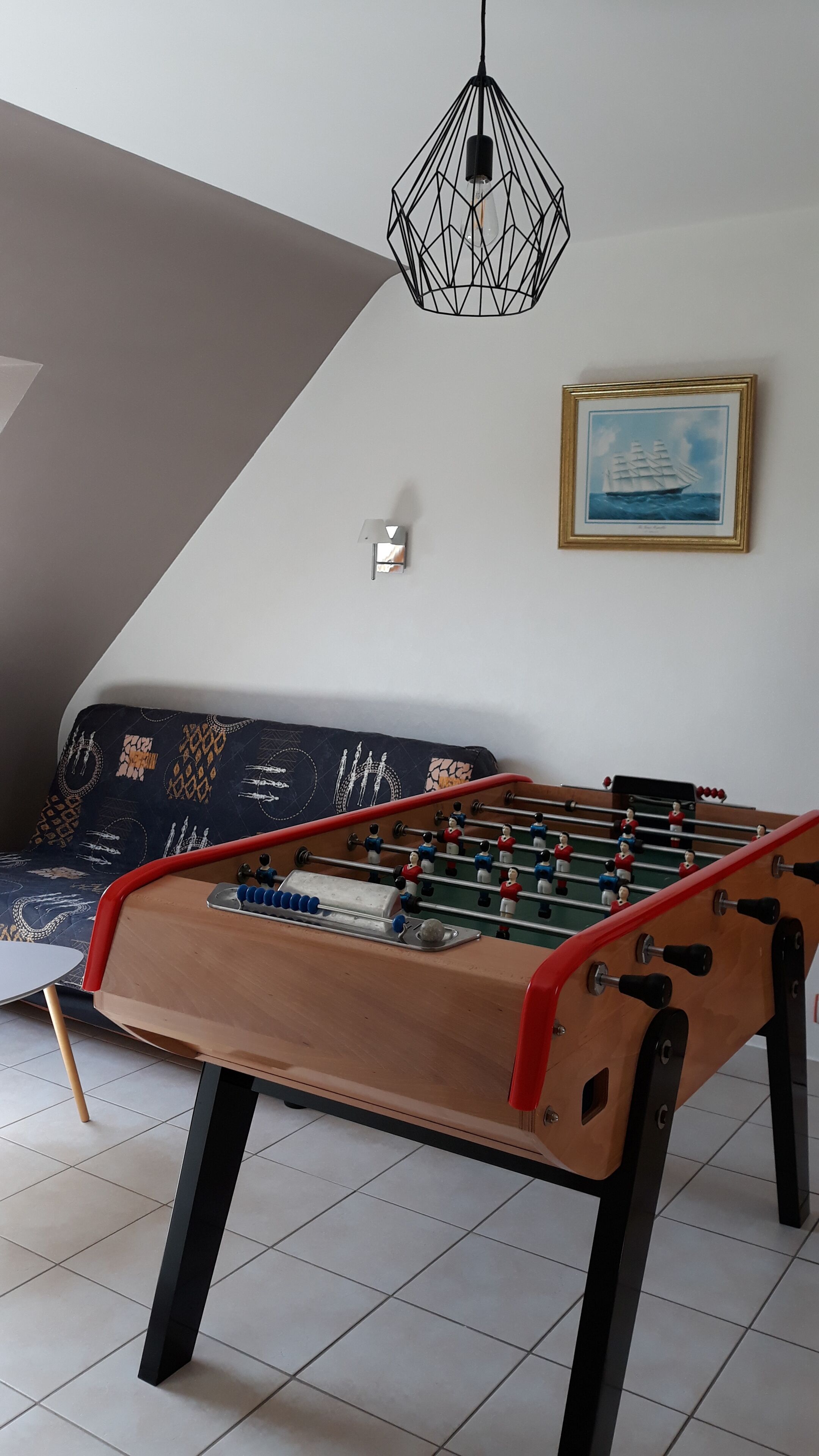 Ein Kicker steht in einem Raum mit einem sofa in geometrischem Muster, einer wandmontierten Lampe und einem gerahmten Bild von einem Schiff.