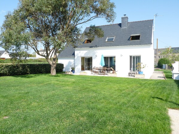 Una casa blanca con techo oscuro, un árbol grande en el jardín y un patio con mobiliario exterior.