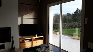 Un salon doté d’un téléviseur à écran plat, d’un meuble de rangement en bois et d’une grande porte-fenêtre coulissante donnant sur une terrasse avec vue sur le jardin.
