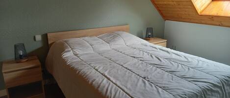 A bedroom with a sloped ceiling, a bed with a white duvet, two bedside tables, and a wall-mounted lamp.