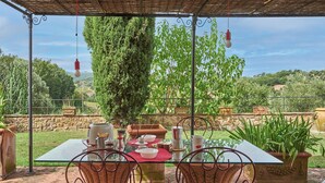 An outdoor dining area with a glass table and wicker chairs, surrounded by greenery and stone walls. - Apartment with terrace and enchanting view (Torrita di Siena)